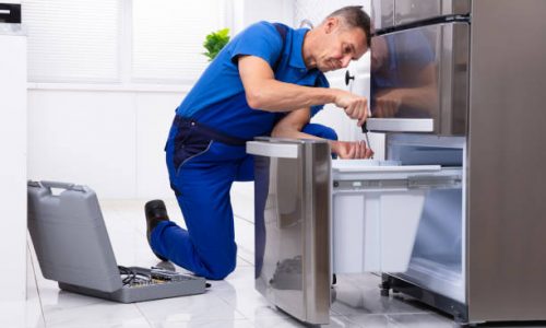 Mature Male Serviceman Repairing Refrigerator With Toolbox In  Kitchen