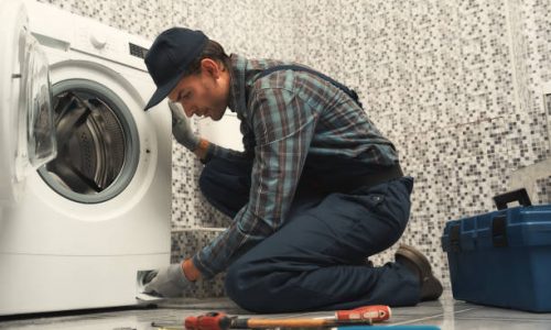 Portrait of young handyman in uniform sitting next to broken washing machine and trying to fix it. Horizontal shot. Side view