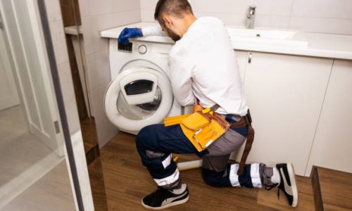 The young handsome repairman in worker is fixing the washing machine in the bathroom.