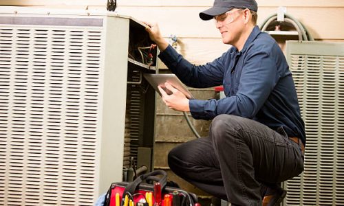 Repairmen works on a home's air conditioner unit outdoors. He is checking the compressor inside the unit using a digital tablet.  He wears a navy blue uniform and his safety glasses.  Tools inside toolbox on ground.